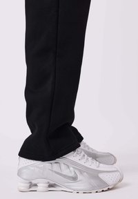 Gambe che indossano pantaloni neri e scarpe da ginnastica Nike Shox bianche con swoosh argentato, mostrate su uno sfondo chiaro e uniforme.