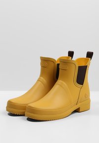 Botas de tobillo impermeables amarillas hechas de goma, con paneles laterales elásticos marrones, suelas texturizadas y un acabado suave.