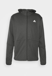 Sudadera de cremallera gris oscuro con una textura suave, capucha ajustada y un logo blanco de adidas en el pecho; cuenta con bolsillos laterales y una elasticidad moderada.