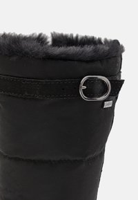 Svart polstret støvel med kunstig pelsfôring på toppen, med sort stropp og detaljer i sølvhardware. Glatt tekstur med sydde sømmer.