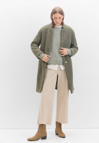Grüner strukturierter Mantel mit hohem Kragen, getragen über einem hellgrauen Strickpullover. Eine weiße, weit geschnittene Hose und braune Stiefeletten vervollständigen den Look.