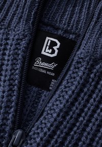 Gros plan d'un tissu tricoté bleu foncé avec une fermeture éclair partiellement ouverte montrant une étiquette noire indiquant Brandit Individual Wear.