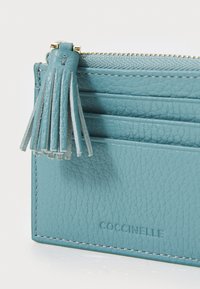 Tirkīza krāsas ādas kartes turētājs ar teksturētu apdari, rāvējslēdzēja aizdari un ādas pušķi. Iekļauj kartes kabatas un izsistus zīmolus.