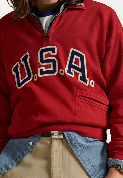 Pulover roșu cu fermoar pe jumătate și inscripția "U.S.A." în crem și bleumarin. Dispune de două buzunare frontale și este purtat peste o cămașă din denim.