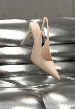 Beež lakknahast kõrge kontsaga slingback-king, millel on silt "VEGAN", lebamas metallik-hõbedasel tassitud pinnal.