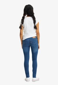 Vaqueros ajustados de mezclilla en color azul, con un lavado claro, bolsillos tradicionales y un parche de cuero marrón en la cintura trasera. Camiseta blanca y zapatillas deportivas.