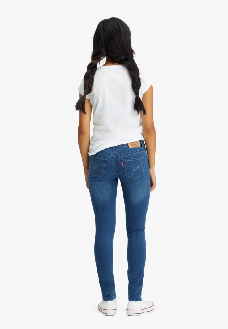 Schmale blaue Jeans aus Denim, mit einem hellen Waschung, traditionellen Taschen und einem hellbraunen Lederpatch am hinteren Bund. Weißes T-Shirt und Turnschuhe.
