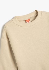Beige sweatshirt med en ribbad crewneck och nerfallna axelsömmar. Slät textur, minimal sömnad, med en liten orange etikett.