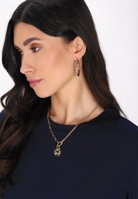 Zlatý řetízek s květinovým přívěskem, doplněný lesklými zlatými kruhovými náušnicemi. Modelka má na sobě námořnickou modrou blůzu a dlouhé tmavé vlasy.