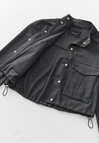Veste boutonnée gris foncé avec une grande poche avant, ourlet à cordon de serrage et col, posée à plat sur une surface blanche.