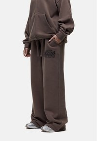 Brauner Fleecepullover mit einer Vordertasche und passenden weiten Hosen mit einem schwarzen Logo-Print. Graue Sneakers mit strukturierten Details.