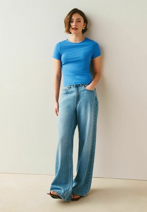 Kvinde iført en blå tætsiddende kortærmet top med vidde bukser i lyseblå, stående mod en ensfarvet lys væg.