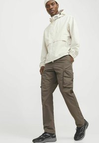 Rompevientos beige claro con cierre de cremallera y puños elásticos, combinado con pantalones cargo marrones con bolsillos laterales. Zapatillas deportivas negras.
