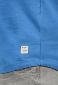 Blå bomulls T-skjorte med en etikett som leser "BLEND GLOBAL JEANMAKER BYGGET I BLÅ" på den nederste kanten, med en jevn tekstur.