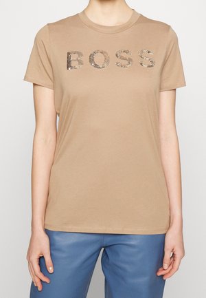 Beżowy T-shirt z krótkim rękawem z cekinowym napisem „BOSS”, noszony z niebieskimi spodniami na osobie z rozluźnionymi ramionami swobodnie opuszczonymi wzdłuż boków.
