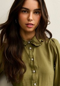 Olijfgroene blouse met een gerimpelde kraag, parelknopen als accenten en lange mouwen met geplooide schouders; zacht, gestructureerd stof.