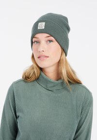 Beanie en tricot côtelé vert avec un revers et une petite étiquette logo. Un haut col roulé côtelé vert assorti complète le beanie.