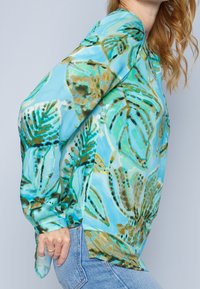 Blusa a maniche lunghe in un tessuto azzurro chiaro con un motivo di foglie tropicali in verde e marrone. Texture seta con dettagli a spacchi laterali.