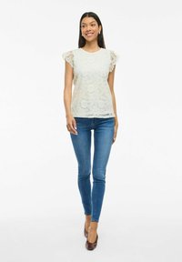 Blonde top i lys creme med korte flæseærmer, parret med tætsiddende blå jeans og brune hæle. Toppen har blomsterprint.