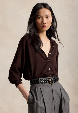Femme aux longs cheveux noirs porte un chemisier marron foncé boutonné, un pantalon gris plissé et une ceinture tressée noire avec une boucle dorée.