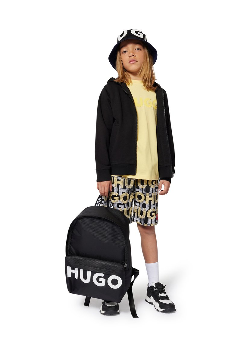 Haut à capuche noir zippé, t-shirt jaune, shorts à motifs et baskets noir et blanc ; tenant un sac à dos noir avec le logo "HUGO" blanc.