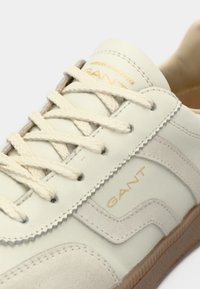 Ténis bege claro feitos de couro e camurça, com atacadores creme, sola de borracha bege e detalhe do logótipo "GANT" dourado na lateral.