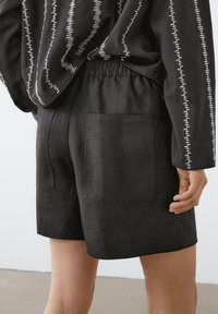 Zwarte shorts met elastische tailleband en grote achterzakken, gedragen met een zwarte top met lange mouwen die verticale witte stikpatronen heeft.