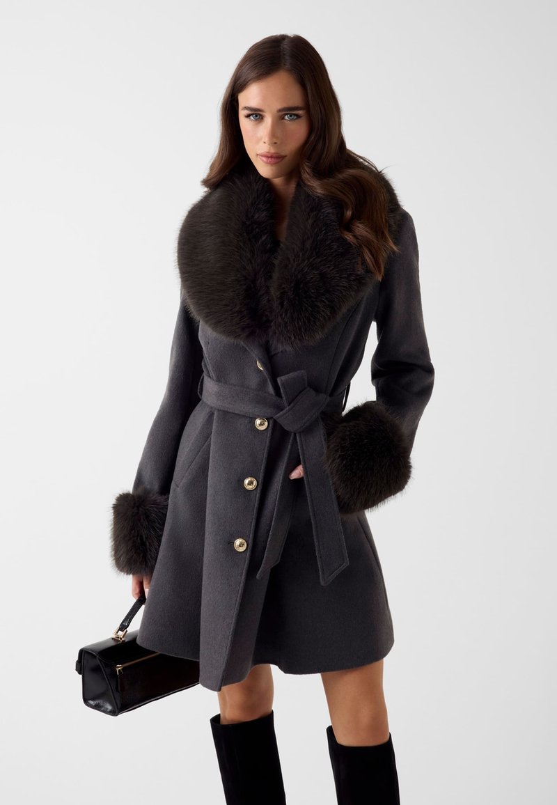 Cappotto grigio di lana con ampio colletto di pelliccia e polsini in pelliccia, caratterizzato da bottoni dorati, vita con cintura e taglio dritto. Borsa nera e stivali al ginocchio.