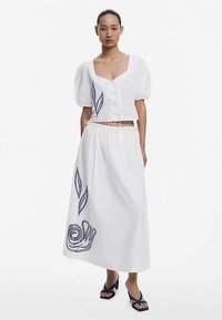 Bijela kratka bluza s naboranim rukavima i gumbima sprijeda, uparen s dugom bijelom suknjom koja ima plave vezenice u obliku lista i spiralnih uzoraka.