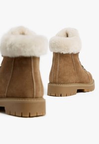 Braune Wildleder-Stiefeletten mit einem weißen Kunstfellbund, ausgestattet mit einer dicken Gummisohle mit tiefen Profilrillen und kontrastierenden weißen Nähten.