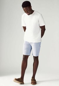 Camiseta blanca de manga corta, pantalones cortos a rayas azul claro, sandalias marrones. Tejido suave, corte relajado, diseño casual.