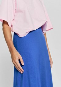 Hellrosa, oversized Kurzarmoberteil, kombiniert mit einem langen blauen Rock. Die Stoffe wirken weich, mit einer glatten Textur und subtiler Drapierung.