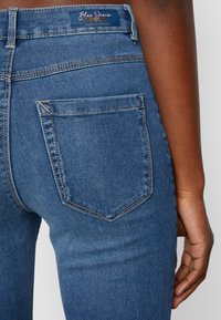 Jeans de ganga azul com um design ajustado, apresentando um bolso traseiro e costuras cosidas. O tecido tem uma textura suave com desbotamento subtil.