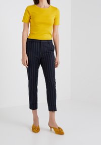 Žlutý přiléhavý top s krátkým rukávem v kombinaci s námořnickými kalhotami s proužkem, ohrnutými u kotníků. Modelka nosí hořčicové lodičky na vysokém podpatku se zdobným mašlí.