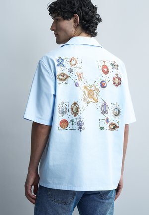 Άνδρας με σγουρά σκούρα μαλλιά, φοράει ανοιχτό μπλε κοντομάνικο πουκάμισο με πολύχρωμα σχέδια σε έμβλημα στην πλάτη, συνδυασμένο με μπλε τζιν.