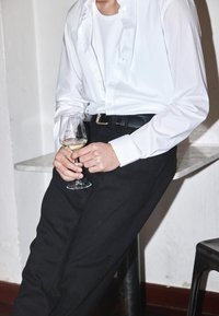 Hombre vestido con una camisa blanca y pantalones negros, sosteniendo una copa de vino blanco mientras se apoya en una encimera de mármol.