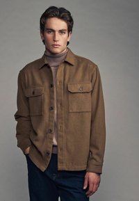 Brun uld skjortejakke med to frontlommer, sorte knapper og en blød tekstur, båret over en lys turtleneck og kombineret med mørke jeans.