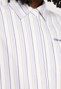 Camicia a righe con strette righe verticali in lavanda, crema e navy su uno sfondo bianco; include una tasca sul petto con logo.