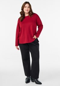 Crvena majica dugih rukava s okruglim izrezom, u kombinaciji s širokim crnim hlačama. Majica ima bočne proreze; oba komada imaju glatku teksturu.