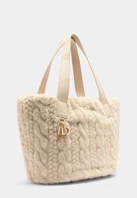 Sac fourre-tout en peluche beige avec une texture tricotée, doté de poignées en cuir lisse et d'un charm en or sur le côté.
