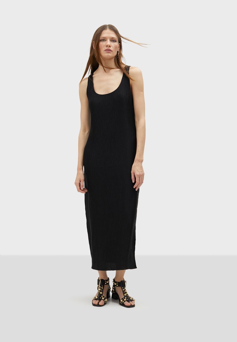 Crna bezrukavna maksi haljina s izrezom u obliku uvale i vertikalnim nabranjima. Uparena s robusnim crnim sandalama s zlatnim akcentima.