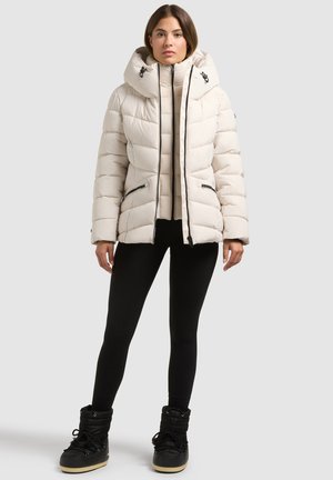 Femme portant une longue doudoune beige, des leggings noirs et des bottes d'hiver noires, debout, les bras détendus, devant un fond gris.