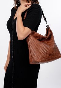 Brązowa torebka hobo z plecionej skóry, o luźnym fasonie, z ciemnym paskiem oraz prostokątną metką marki z przodu.