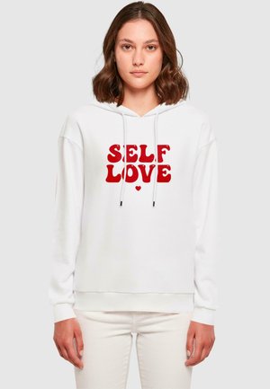 Biała bluza z kapturem z napisem "SELF LOVE" w pogrubionej czerwonej literze. Ma luźny krój, sznurki i ściągacze przy rękawach. Gładka faktura.
