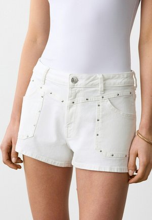 Pantaloncini di jeans bianchi con borchie argentate indossati da una persona con una maglia bianca, che mostra dalla vita a metà coscia, con le mani rilassate ai lati.