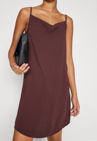 Mörkröd ärmlös klänning med cowl-krage, tillverkad av strukturerat tyg, med en avslappnad passform. Modellen håller en svart kuvertväska.