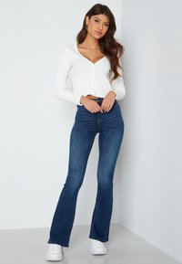 Witte geribbelde langekleed cropped cardigan met knopen aan de voorkant, gedragen met donkerblauwe flair jeans en witte sneakers, staande tegen een lichte achtergrond.