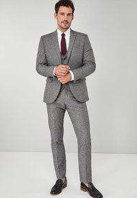 Costum gri din țesătură tweed, cu o croială ajustată, având rever în formă de notch, închidere cu trei nasturi și pantaloni cu față plată. Asortat cu o cămașă albă și o cravată vișinie.