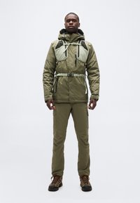 Άνδρας που στέκεται φορώντας πλήρη εξοπλισμό υπαίθρου σε ελαιώδες πράσινο, συμπεριλαμβανομένου μπουφάν, παντελόνι καρό, παπούτσια πεζοπορίας και θώρακα με ιμάντες και πόρπες.