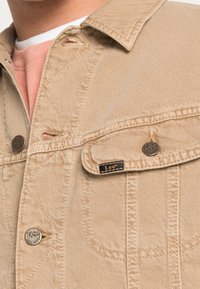 Beige farkkutakki, jossa on nappikiinnitys edessä, läppätaskut ja mustat brändiakkesennat. Ominaisuuksia ovat teksturoitu pinta ja ommellut yksityiskohdat.